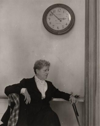 [Woman sitting at a table, under a large clock]