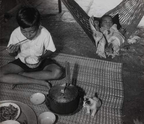 Boy having a meal with baby in a hammock next to him, Vietnam