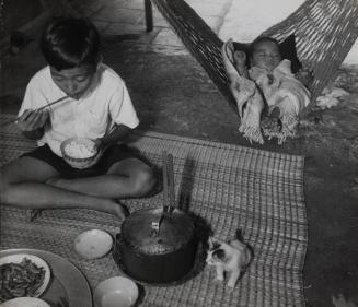 Boy having a meal with baby in a hammock next to him, Vietnam