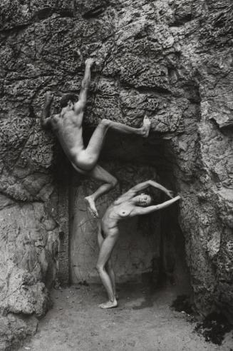 A nude man hangs on a rock wall above an arching, nude Kate