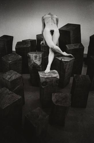 Kate, nude, bends over several carved logs indoors, USA