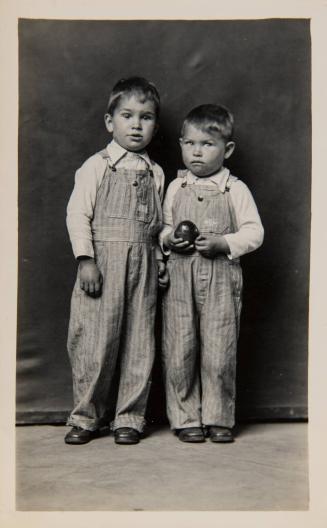 [Two standing boys in denim overalls, one holding an apple]