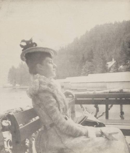 [Woman in hat and dress sits on bench]