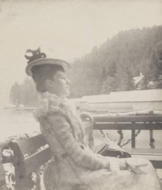[Woman in hat and dress sits on bench]