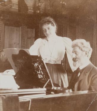 [Woman stands next to seated man at piano]