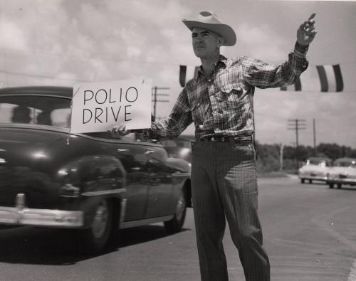 [Man standing in the middle of the street holding a sign that says "Polio Drive"]